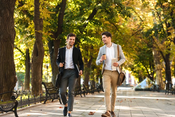 bearded businessmen in suits walking outdoor through green park with takeaway coffee and laptop, during sunny day