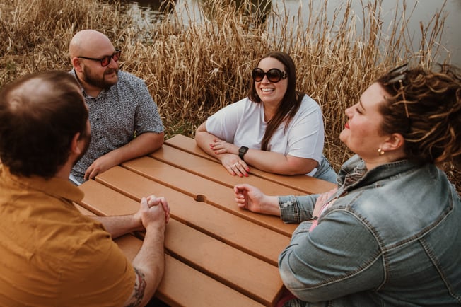 4 friends sit around table laughing - AdobeStock_603882870 - June 2024 Blog