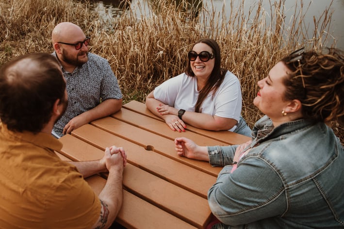 4 friends sit around table laughing - AdobeStock_603882870 - June 2024 Blog