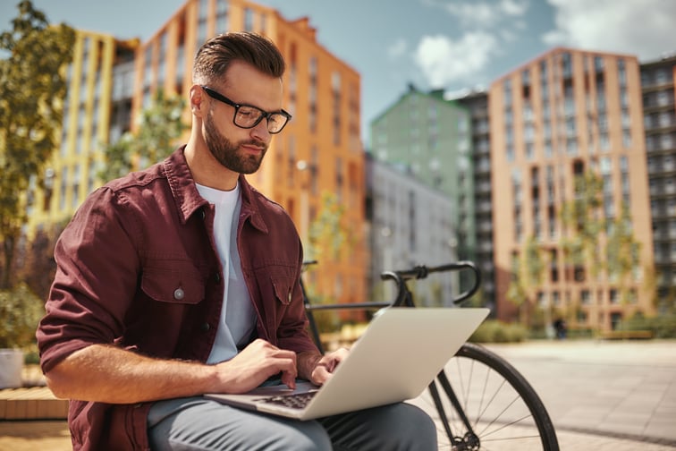 man working on laptop while sitting on the bench near his bicycle - AdobeStock_283083147 - June 2024 Blog